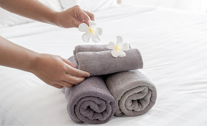 Three rolled gray towels are stacked on a white surface with two white flowers placed on top while a person arranges one of the flowers. Description generated by AI