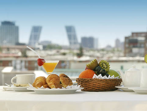 A breakfast setting on a table includes a cup of coffee, croissants on a plate, a cocktail with a strawberry garnish, a basket of assorted fruits, and a teapot with a cityscape in the background. Description generated by AI