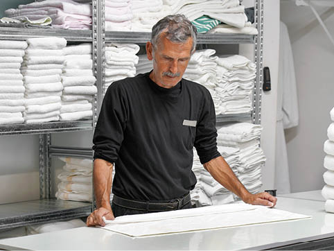 A senior person wearing a black long-sleeve shirt is folding white towels in a laundry room with shelves stacked with neatly folded towels. Description generated by AI