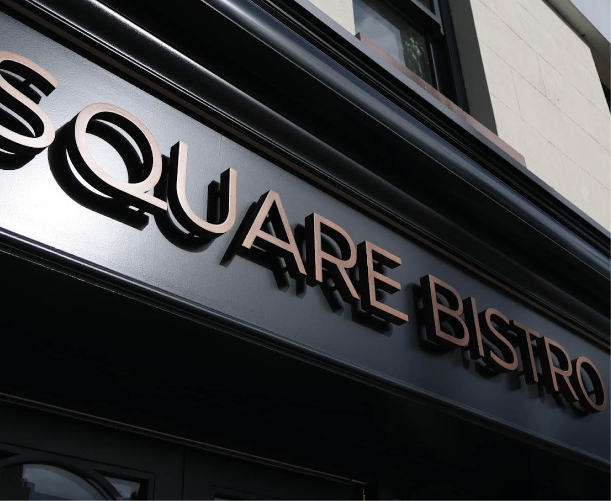 A close-up view of a black sign with raised bronze letters spelling “SQUARE BISTRO" mounted above a building entrance. Description generated by AI
