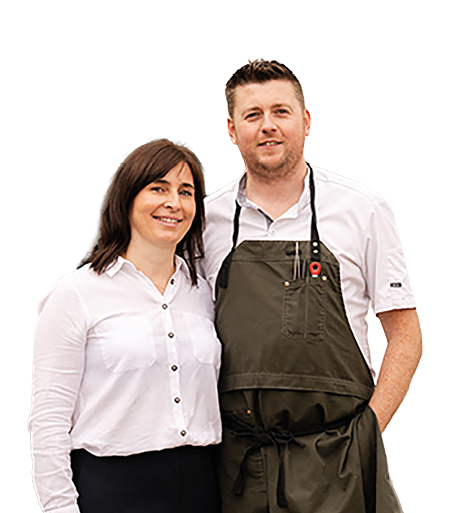 Two adults stand close together smiling, one wearing a white button-up shirt and the other wearing a white shirt with a dark apron. Description generated by AI