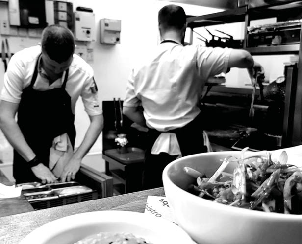 Two adults wearing aprons are working in a kitchen with one preparing food on a counter and the other cooking at an oven, while a bowl of food is in the foreground. Description generated by AI