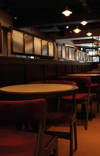 A dimly lit restaurant interior with round wooden tables and red cushioned chairs arranged in rows along a wooden partition with hanging pendant lights above. Description generated by AI