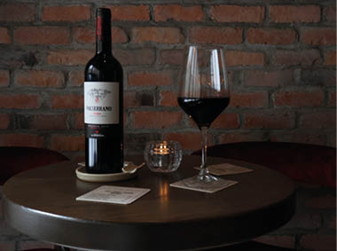 A bottle of red wine, a filled wine glass, a lit candle, and two coasters are arranged on a round wooden table against a brick wall background. Description generated by AI