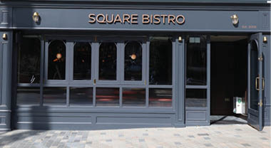 A dark gray storefront with large windows and a door slightly open displays the name Square Bistro in gold letters above the entrance. Description generated by AI
