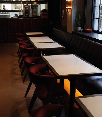A dimly lit restaurant interior with a row of empty tables and red cushioned chairs along a dark bench seating near a window. Description generated by AI