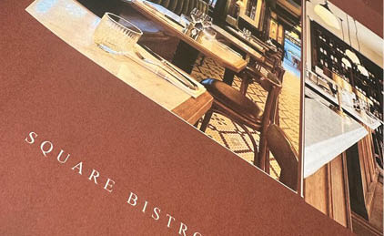 A cozy bistro interior with wooden tables and chairs set with glassware and cutlery, featuring warm lighting and a patterned carpet. Description generated by AI