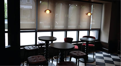 A dimly lit caf interior features round tables with cushioned stools arranged near large windows covered by partially lowered blinds and two hanging pendant lights.  Description generated by AI
