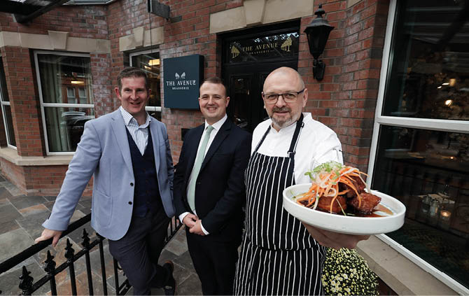 Three adults dressed in formal and chef attire stand smiling outside a restaurant named The Avenue Brasserie, with one holding a plate of food. Description generated by AI