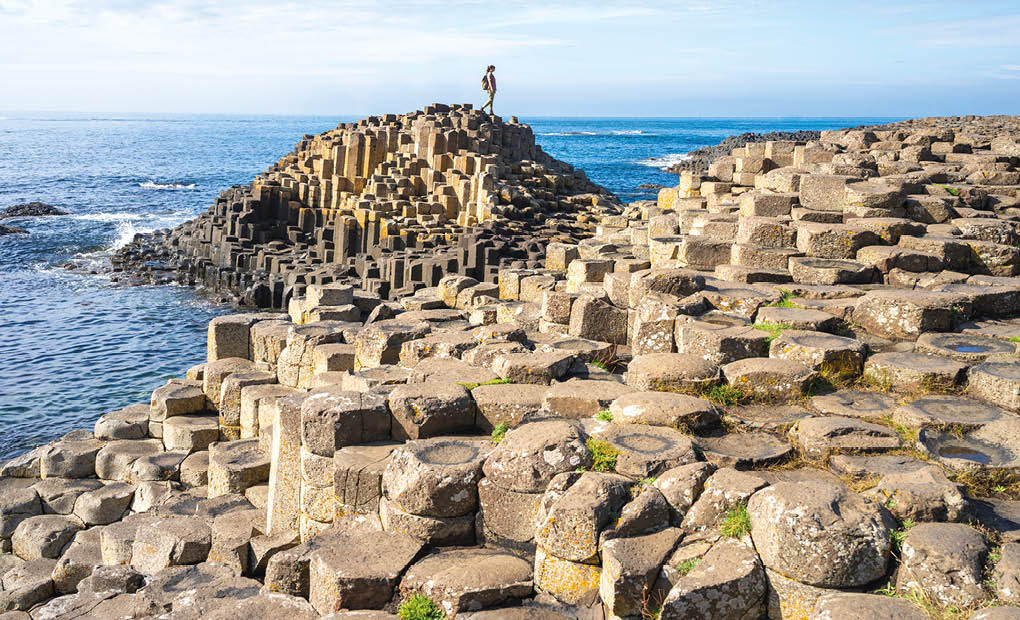 A person stands on top of a natural formation of hexagonal basalt columns by the ocean under a partly cloudy sky. Description generated by AI