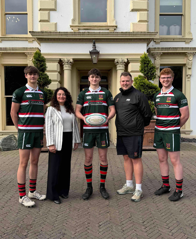 Five people stand in front of a large building, three wearing matching green sports uniforms with red and white stripes, one holding a rugby ball, and two wearing casual clothing. Description generated by AI