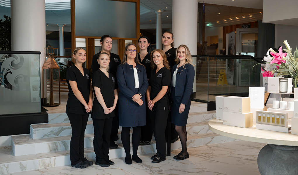 A group of eight adults dressed in professional black and navy attire stand together indoors on marble flooring near a table with white boxes and pink flowers. Description generated by AI