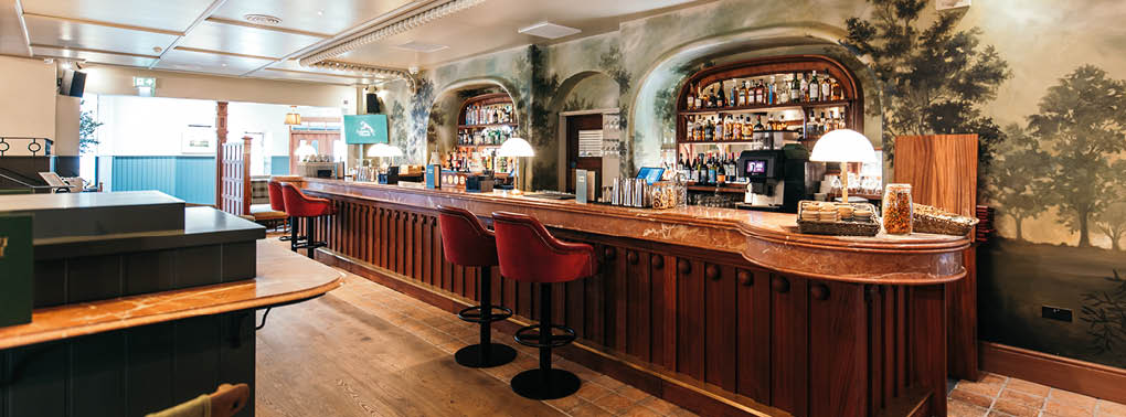 A cozy bar interior with a long wooden counter, red cushioned stools, shelves stocked with bottles, and warm ambient lighting. Description generated by AI