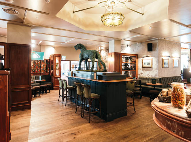 A warmly lit, stylish bar interior features wooden floors, a central counter with high chairs, a large decorative sculpture of an animal on the counter, and cozy seating areas with framed artwork on the walls. Description generated by AI