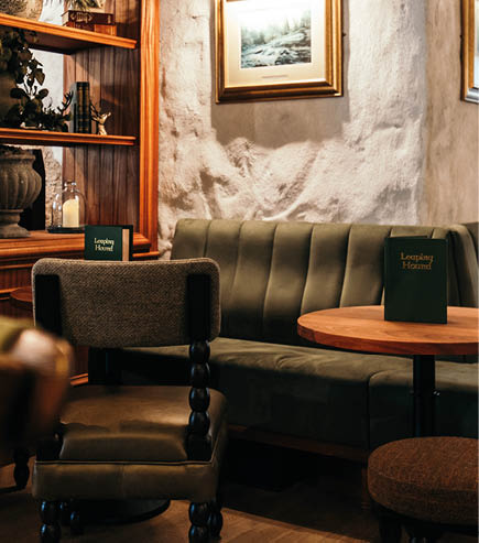 A cozy seating area with a green upholstered bench, wooden table, cushioned chairs, a framed landscape painting on a textured wall, and a wooden shelf with decorative plants and objects. Description generated by AI