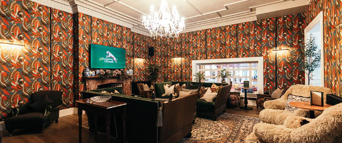 A cozy living room features patterned wallpaper, a chandelier, a wall-mounted TV, dark furniture, a large window, and a fluffy armchair with a side table and plant. Description generated by AI