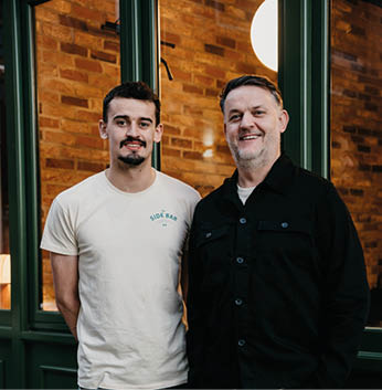 Two adults stand side by side smiling in front of a brick wall and large windows, one wearing a light-colored t-shirt and the other wearing a black button-up shirt. Description generated by AI