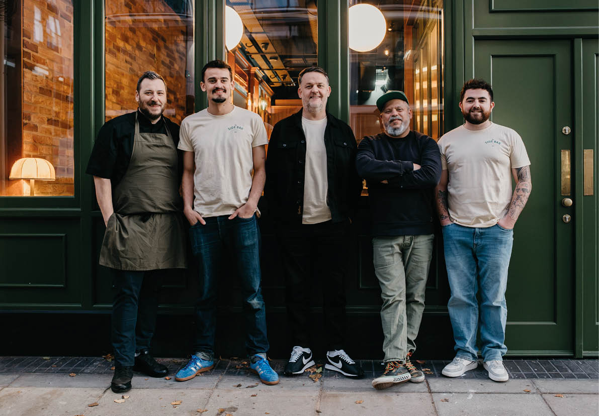Five adults stand side by side outside a building with green doors and windows, dressed casually with some wearing aprons and others in t-shirts and jeans. Description generated by AI