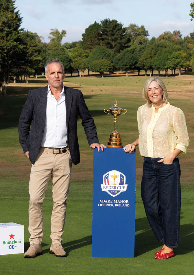 Two adults stand on a golf course next to a blue pedestal holding a trophy, with two white boxes featuring a Heineken logo placed on the grass nearby. Description generated by AI