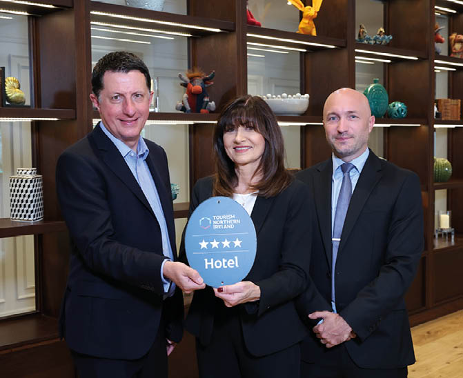 Three adults dressed in business attire stand indoors holding a blue plaque with four stars and the word Hotel, smiling in front of wooden shelves with decorative items. Description generated by AI
