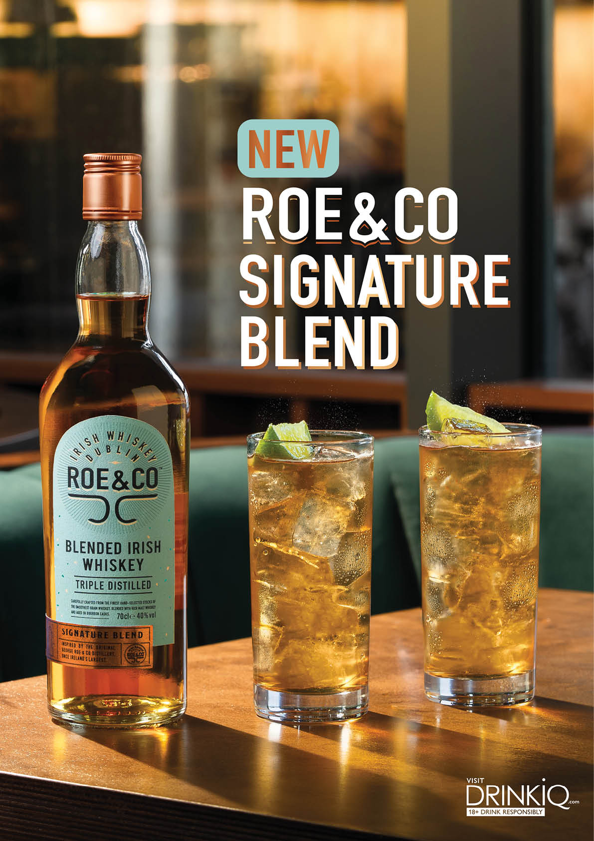A bottle of Roe & Co blended Irish whiskey is displayed on a wooden table alongside two tall glasses filled with whiskey and ice, garnished with lime wedges, set against a blurred background. Description generated by AI