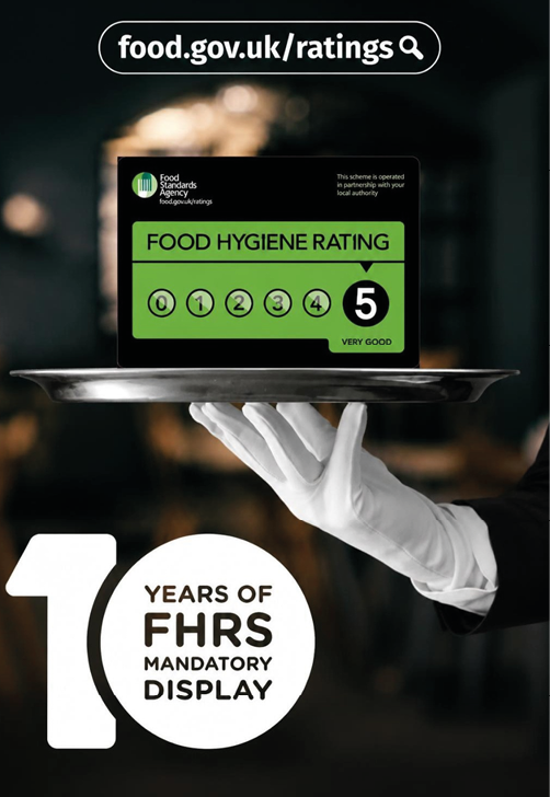 A gloved hand holds a silver tray displaying a food hygiene rating card with the highest rating and a graphic celebrating ten years of mandatory food hygiene rating display. Description generated by AI