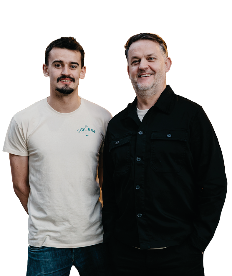Two adults stand side by side smiling with one wearing a light-colored t-shirt and jeans and the other wearing a black button-up shirt and dark pants. Description generated by AI