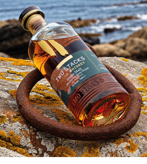 A bottle of Two Stacks Single Malt Irish Whiskey is resting on a rusty metal ring on a rocky surface with a blurred ocean background. Description generated by AI