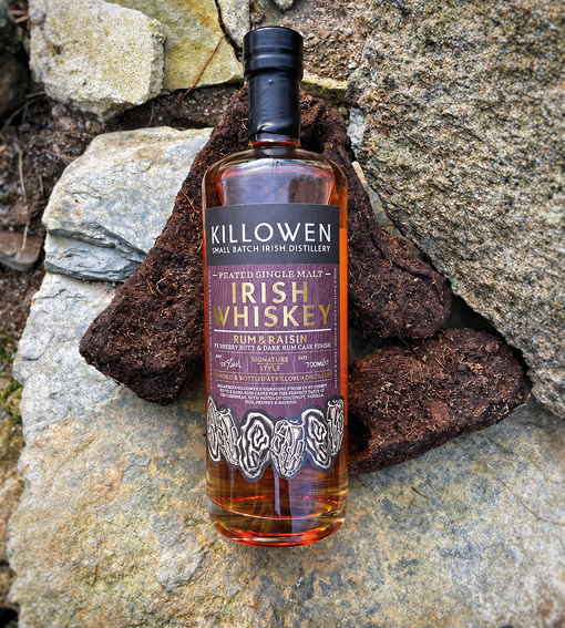 A bottle of Killowen Irish Whiskey is placed upright on a large stone with smaller rocks in the background. Description generated by AI