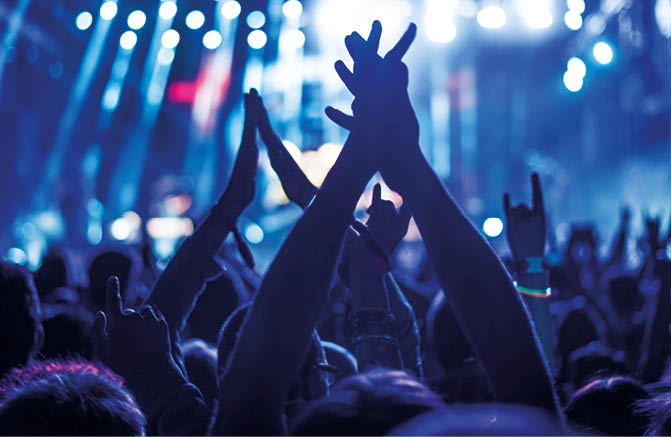 Audience with hands raised at a music festival and lights streaming down from above the stage. Soft focus, blurred movement.