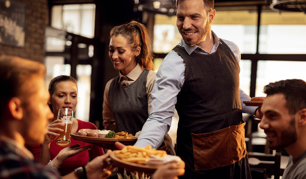 Happy waiters bringing food at the table and serving group of friends in a restaurant.