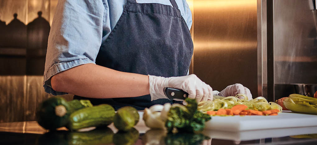Calm and focused female chef standing in a dark kitchen next to cutting board while cutting vegetables on it, wearing apron and denim shirt, posing for the camera, cooking show look