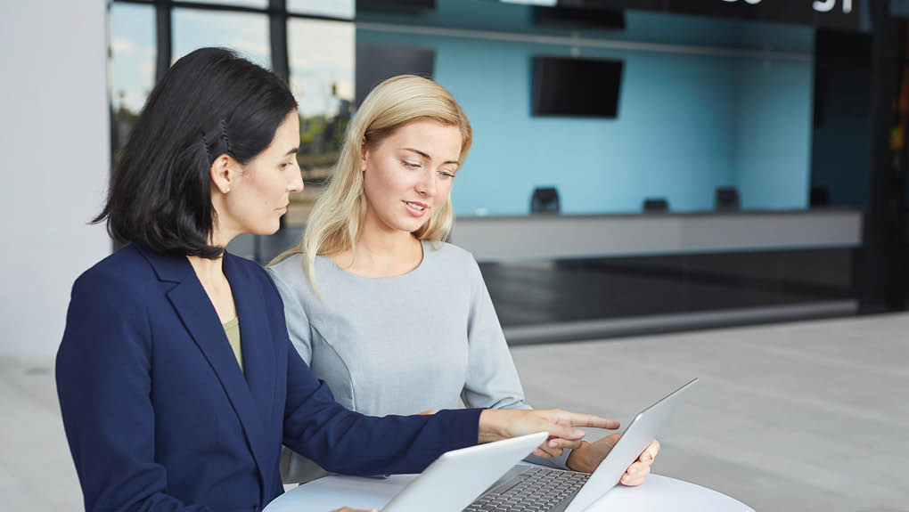 Waist up portrait of two successful businesswomen discussing work while standing at desk in office building and using laptop, copy space