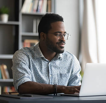 Satisfied African American man wearing glasses looking at computer screen, reading good news in email, chatting in social network with friends, freelancer blogger working on online project