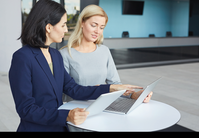 Waist up portrait of two successful businesswomen discussing work while standing at desk in office building and using laptop, copy space