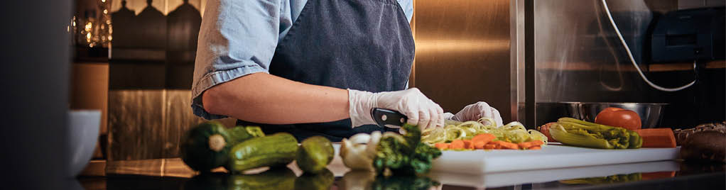Calm and focused female chef standing in a dark kitchen next to cutting board while cutting vegetables on it, wearing apron and denim shirt, posing for the camera, cooking show look