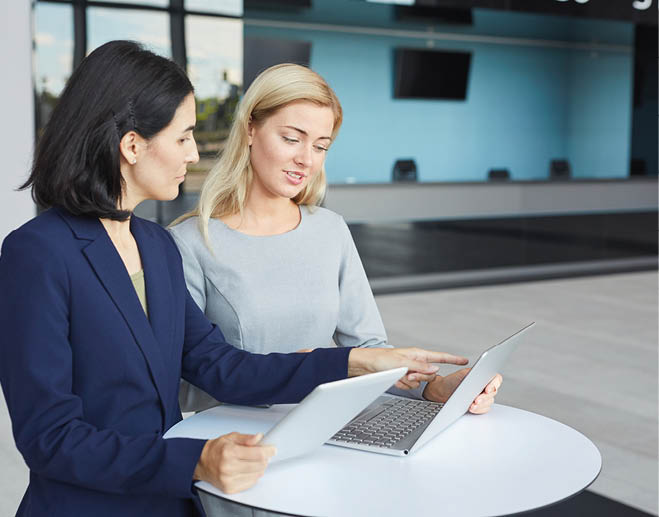 Waist up portrait of two successful businesswomen discussing work while standing at desk in office building and using laptop, copy space