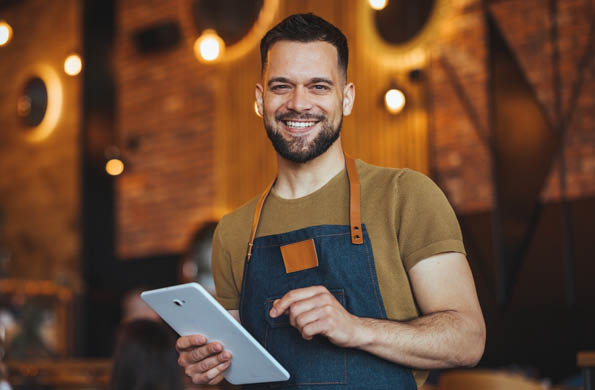 Happy waiter in denim apron holding a tablet, smiling warmly in a trendy restaurant with stylish decor and ambient lighting. Ideal for concepts of hospitality and customer service.