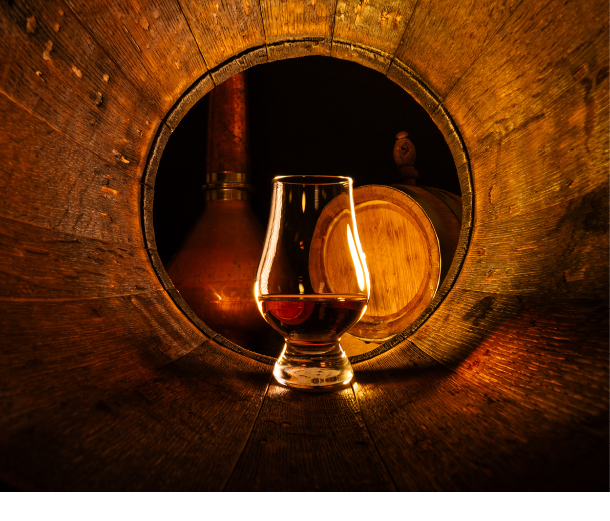 A glass of scotch whiskey in old oak barrel. Copper alambic and small barrel on background. Traditional alcohol distillery concept