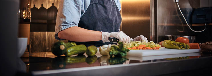 Calm and focused female chef standing in a dark kitchen next to cutting board while cutting vegetables on it, wearing apron and denim shirt, posing for the camera, cooking show look